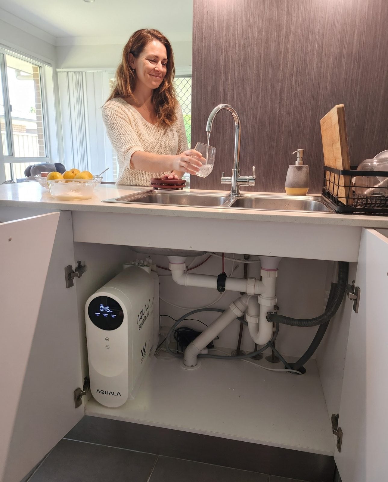 A woman pours a glass of water at her kitchen bench. In the open cupboard below, a P500 stands beside the pipes.