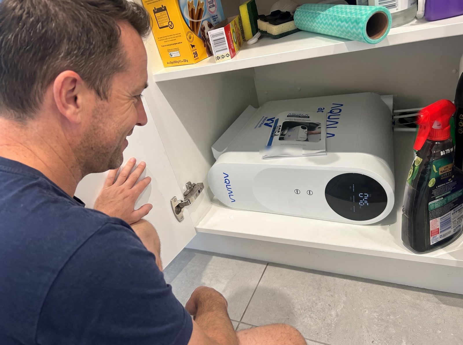 A man crouches at an open cupboard door. The P500 lies on its side on the shelf with its manual on top.