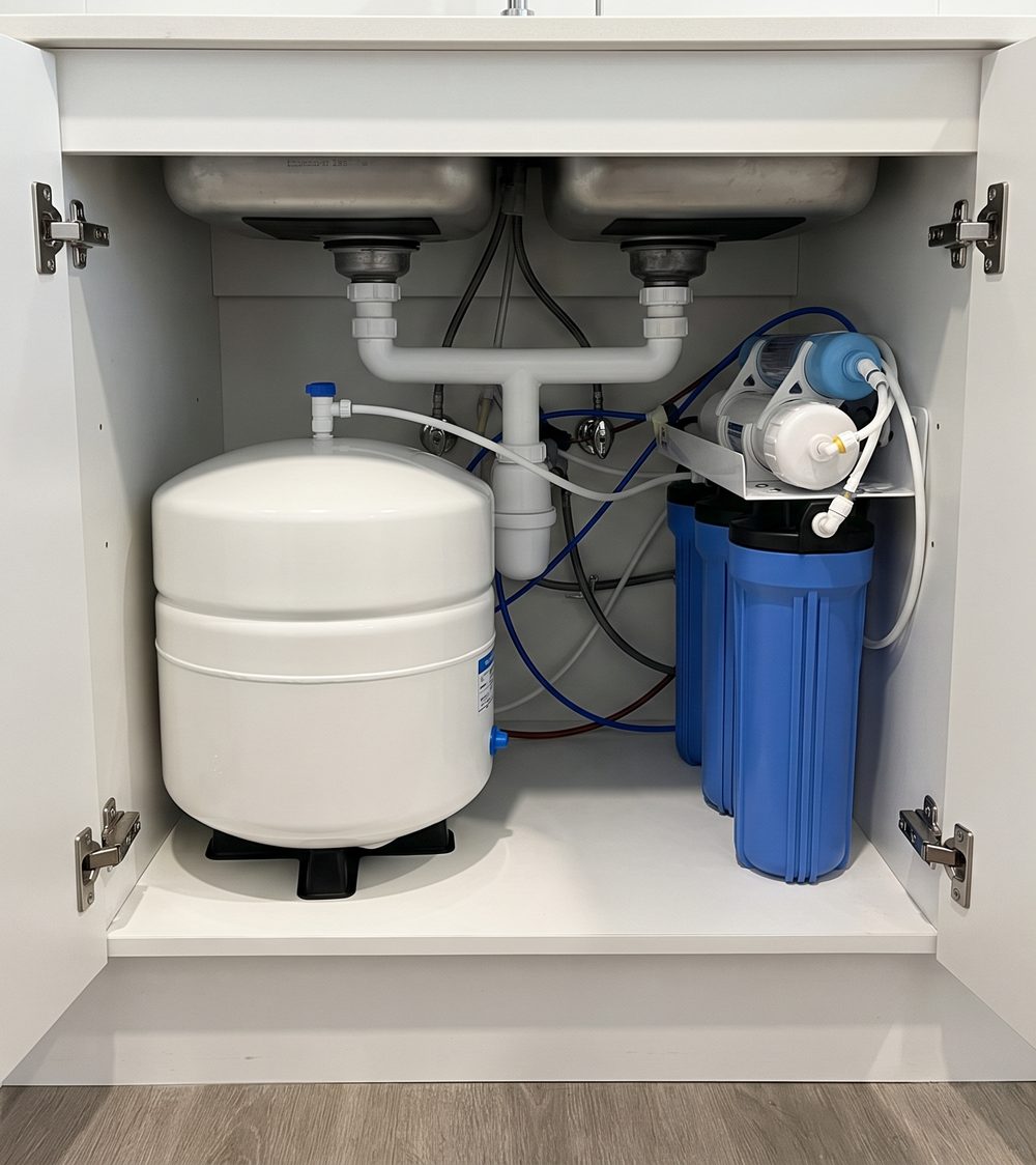 Under a double sink, a tank system: a white storage tank beside blue filter housings and tubing.
